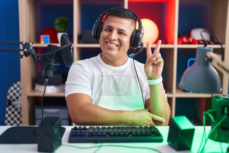 Photo for Young hispanic man playing video games smiling with happy face winking at the camera doing victory sign with fingers. number two. - Royalty Free Image