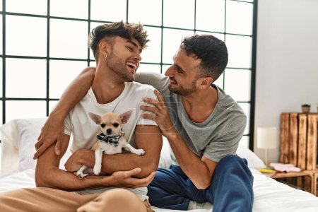 Téléchargez les photos : Deux hommes hispaniques couple s'embrassant assis sur le lit avec chihuahua dans la chambre - en image libre de droit