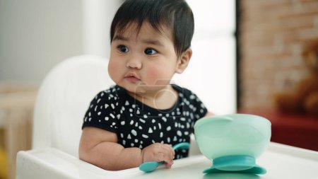 Photo for Adorable hispanic baby sitting on highchair holding spoon at home - Royalty Free Image