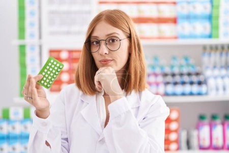 Téléchargez les photos : Young redhead woman working at pharmacy drugstore holding birth control pills serious face thinking about question with hand on chin, thoughtful about confusing idea - en image libre de droit