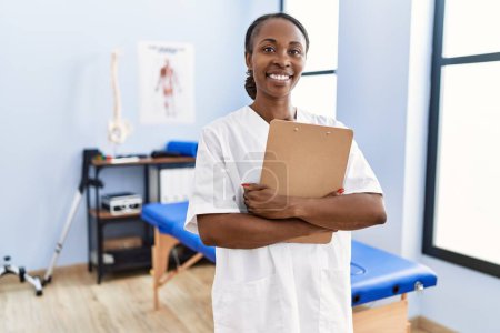 Photo for African american woman physiotherapist smiling confident holding medical report at rehab clinic - Royalty Free Image