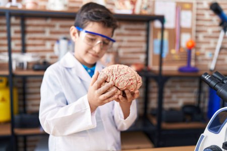 Téléchargez les photos : Adorable garçon hispanique étudiant souriant confiant tenant le cerveau dans la salle de classe du laboratoire - en image libre de droit