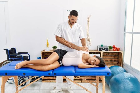 Photo for Latin man and woman wearing physiotherapist uniform having pregnancy rehab session stretching arm at physiotherapy clinic - Royalty Free Image