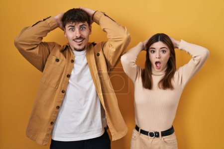 Foto de Young hispanic couple standing over yellow background crazy and scared with hands on head, afraid and surprised of shock with open mouth - Imagen libre de derechos