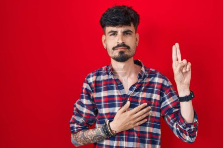 Téléchargez les photos : Jeune homme hispanique avec la barbe debout sur fond rouge souriant jurant avec la main sur la poitrine et les doigts vers le haut, faisant un serment de fidélité promesse - en image libre de droit