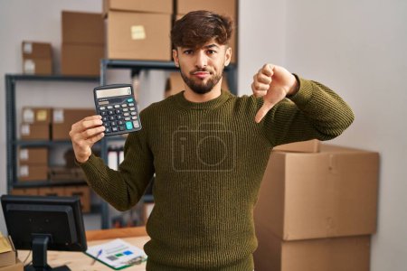 Photo for Arab man with beard working at small business ecommerce holding calculator with angry face, negative sign showing dislike with thumbs down, rejection concept - Royalty Free Image