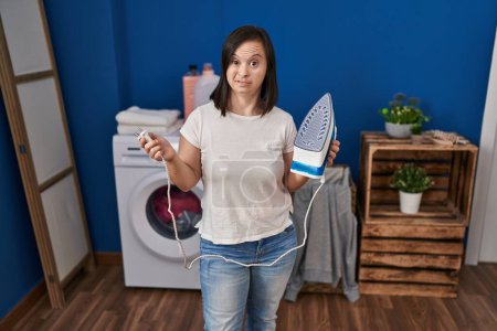 Téléchargez les photos : Hispanic girl with down syndrome ironing clothes at home smiling looking to the side and staring away thinking. - en image libre de droit