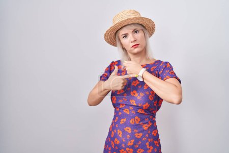 Téléchargez les photos : Young caucasian woman wearing flowers dress and summer hat in hurry pointing to watch time, impatience, looking at the camera with relaxed expression - en image libre de droit