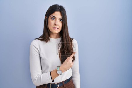 Photo for Young brunette woman standing over blue background pointing aside worried and nervous with forefinger, concerned and surprised expression - Royalty Free Image