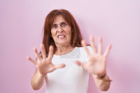 Foto de Mujer de mediana edad de pie sobre el fondo rosa miedo y aterrorizado con la expresión de miedo detener el gesto con las manos, gritando en shock. concepto de pánico. - Imagen libre de derechos