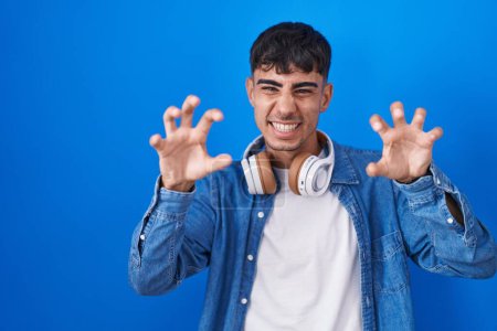 Téléchargez les photos : Jeune homme hispanique debout sur fond bleu souriant drôle faisant griffe geste comme chat, expression agressive et sexy - en image libre de droit