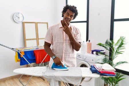 Foto de African man with curly hair ironing clothes at home with hand on chin thinking about question, pensive expression. smiling with thoughtful face. doubt concept. - Imagen libre de derechos