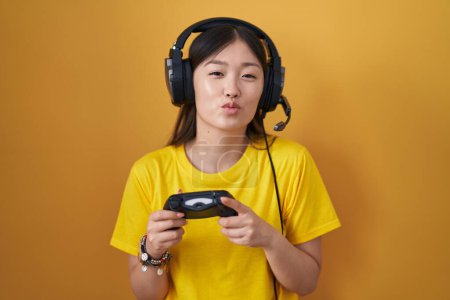 Téléchargez les photos : Chinese young woman playing video game holding controller looking at the camera blowing a kiss on air being lovely and sexy. love expression. - en image libre de droit