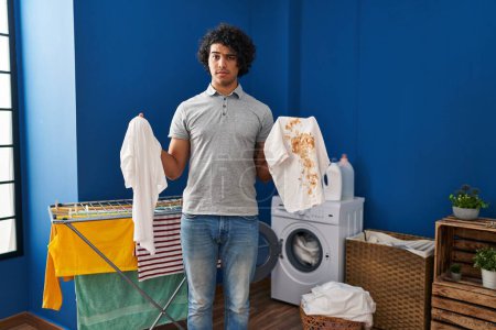Photo for Hispanic man with curly hair holding clean white t shirt and t shirt with dirty stain skeptic and nervous, frowning upset because of problem. negative person. - Royalty Free Image