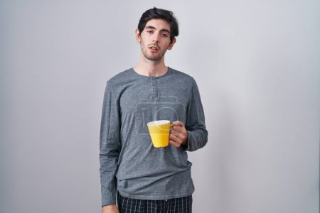 Photo for Young hispanic man wearing pajama drinking a cup of coffee looking sleepy and tired, exhausted for fatigue and hangover, lazy eyes in the morning. - Royalty Free Image