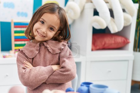 Photo for Adorable hispanic girl smiling confident sitting on table with arms crossed gesture at kindergarten - Royalty Free Image