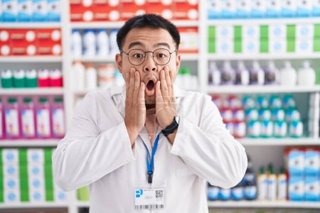 Photo pour Chinese young man working at pharmacy drugstore afraid and shocked, surprise and amazed expression with hands on face - image libre de droit