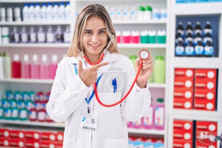 Foto de Young blonde woman working at pharmacy drugstore holding stethoscope smiling happy pointing with hand and finger - Imagen libre de derechos