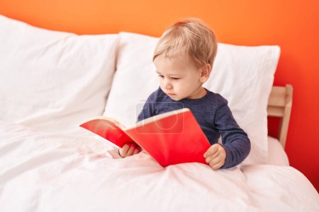 Téléchargez les photos : Adorable tout-petit blond lisant livre d'histoire assis sur le lit dans la chambre - en image libre de droit