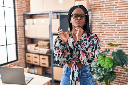 Foto de African woman with braids working at small business ecommerce showing smartphone screen serious face thinking about question with hand on chin, thoughtful about confusing idea - Imagen libre de derechos