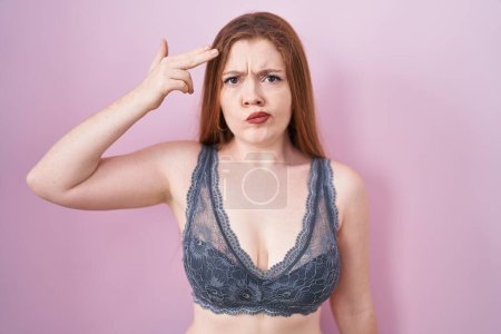 Foto de Redhead woman wearing lingerie over pink background shooting and killing oneself pointing hand and fingers to head like gun, suicide gesture. - Imagen libre de derechos