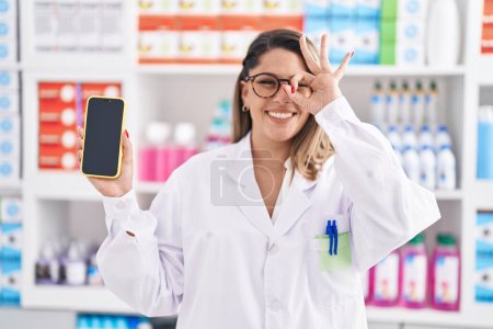 Foto de Blonde woman working at pharmacy drugstore showing smartphone screen smiling happy doing ok sign with hand on eye looking through fingers - Imagen libre de derechos