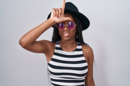 Photo for Young african american with braids wearing hat and sunglasses making fun of people with fingers on forehead doing loser gesture mocking and insulting. - Royalty Free Image
