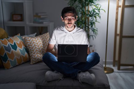Photo for Young hispanic man using laptop at home at night looking at the camera blowing a kiss on air being lovely and sexy. love expression. - Royalty Free Image