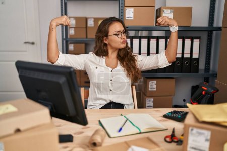 Téléchargez les photos : Young hispanic woman working at small business ecommerce showing arms muscles smiling proud. fitness concept. - en image libre de droit