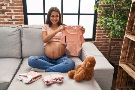 Photo for Young latin woman pregnant holding baby clothes at home - Royalty Free Image