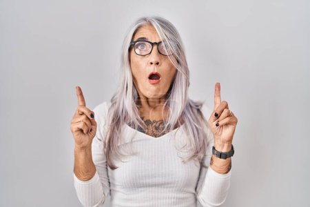 Téléchargez les photos : Middle age woman with grey hair standing over white background amazed and surprised looking up and pointing with fingers and raised arms. - en image libre de droit