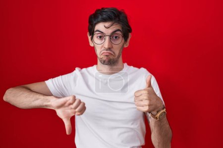 Photo for Young hispanic man standing over red background doing thumbs up and down, disagreement and agreement expression. crazy conflict - Royalty Free Image