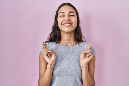 Téléchargez les photos : Young brazilian woman wearing casual t shirt over pink background gesturing finger crossed smiling with hope and eyes closed. luck and superstitious concept. - en image libre de droit