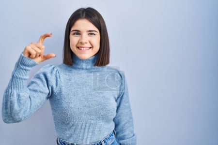 Téléchargez les photos : Jeune femme hispanique debout sur fond bleu geste souriant et confiant avec la main faisant signe de petite taille avec les doigts regardant et la caméra. concept de mesure. - en image libre de droit