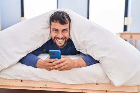 Foto de Hombre hispano joven usando teléfono inteligente acostado en la cama en el dormitorio - Imagen libre de derechos
