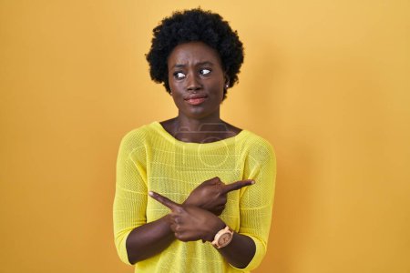Téléchargez les photos : Jeune femme africaine debout sur le studio jaune pointant des deux côtés avec les doigts, direction différente en désaccord - en image libre de droit