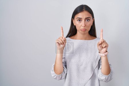 Foto de Young hispanic woman standing over white background pointing up looking sad and upset, indicating direction with fingers, unhappy and depressed. - Imagen libre de derechos