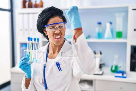 Foto de Middle age hispanic woman working at scientist laboratory annoyed and frustrated shouting with anger, yelling crazy with anger and hand raised - Imagen libre de derechos