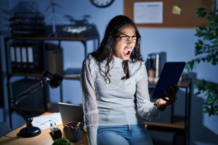 Photo for Young brazilian woman using touchpad at night working at the office angry and mad screaming frustrated and furious, shouting with anger. rage and aggressive concept. - Royalty Free Image