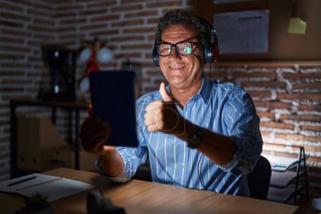 Foto de Middle age hispanic man using touchpad sitting on the table at night approving doing positive gesture with hand, thumbs up smiling and happy for success. winner gesture. - Imagen libre de derechos