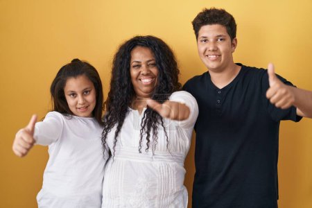 Foto de Family of mother, daughter and son standing over yellow background approving doing positive gesture with hand, thumbs up smiling and happy for success. winner gesture. - Imagen libre de derechos