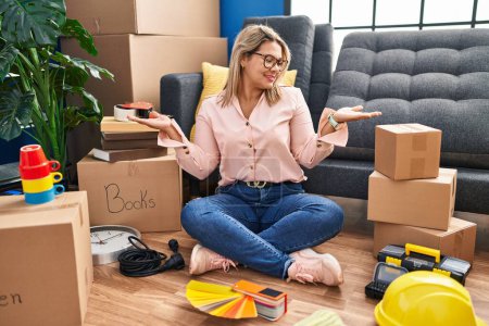 Photo for Young hispanic woman moving to a new home sitting on the floor smiling showing both hands open palms, presenting and advertising comparison and balance - Royalty Free Image
