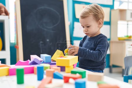 Foto de Adorable niño rubio jugando con bloques de geometría sentado en la mesa en el jardín de infantes - Imagen libre de derechos