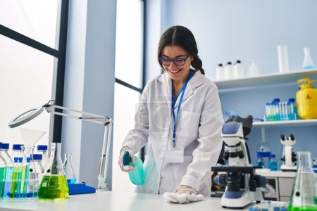 Téléchargez les photos : Jeune hispanique femme portant scientifique uniforme nettoyage à l'aide de gel désinfectant au laboratoire - en image libre de droit