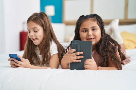 Photo for Two kids using smartphone and touchpad lying on bed at bedroom - Royalty Free Image