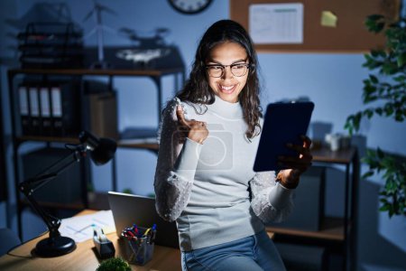 Foto de Mujer brasileña joven usando touchpad en la noche trabajando en la oficina mirando confiado con sonrisa en la cara, señalándose con los dedos orgullosos y felices. - Imagen libre de derechos