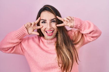 Photo for Young hispanic woman standing over pink background doing peace symbol with fingers over face, smiling cheerful showing victory - Royalty Free Image