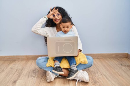 Foto de Joven madre y niño hispano usando computadora portátil sentada en el suelo haciendo buen gesto con la mano sonriente, los ojos mirando a través de los dedos con cara feliz. - Imagen libre de derechos