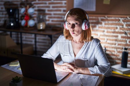 Téléchargez les photos : Young redhead woman working at the office at night wearing headphones making fish face with mouth and squinting eyes, crazy and comical. - en image libre de droit