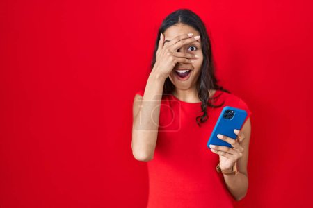 Foto de Young brazilian woman using smartphone over red background peeking in shock covering face and eyes with hand, looking through fingers with embarrassed expression. - Imagen libre de derechos
